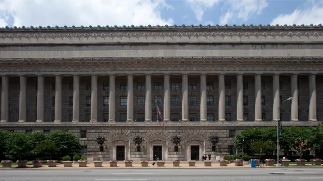 The Herbert C. Hoover Building, headquarters of the U.S. Department of Commerce in Washington, D.C., a long limestone block with flags along the roofline, photographed in 2009. Photo by Carol M. Highsmith, Library of Congress, public domain, via Wikimedia Commons.