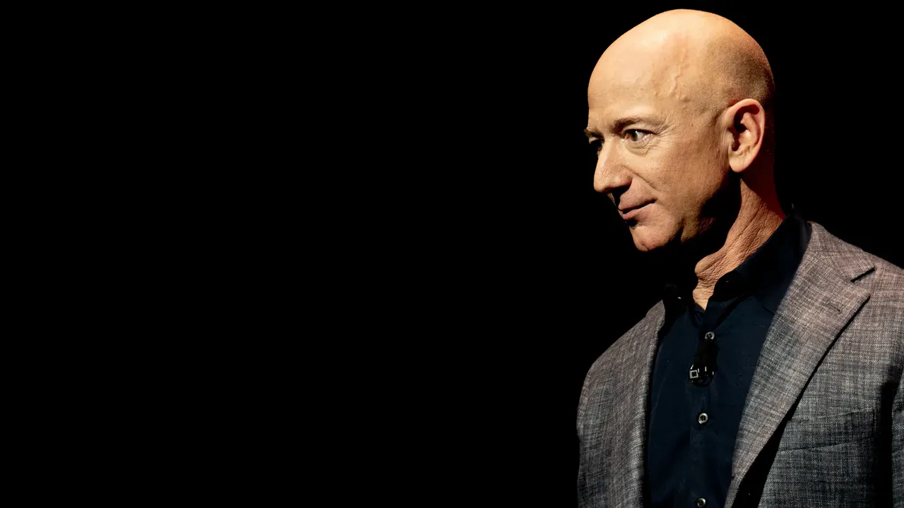 Jeff Bezos, in a dark jacket over a light blue shirt, stands at left beside Blue Origin's prototype lunar lander during a company event in Washington, D.C., on 9 May 2019. The white lander fills the right of the frame, with a Blue Origin banner behind him. The lead image is a 1280 by 720 WebP cropped from that photograph. It is not the 7 October 2026 interview at Cape Canaveral. Photo by Daniel Oberhaus, CC BY 4.0, via Wikimedia Commons.